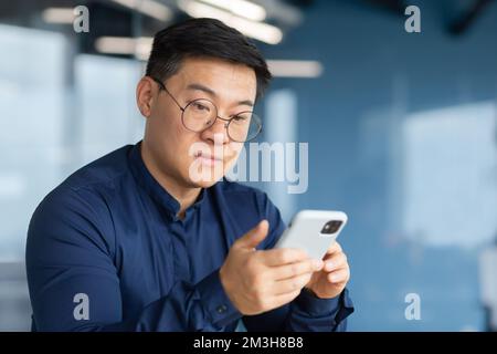 Gros plan d'un homme d'affaires asiatique sérieux et pensif utilisant le téléphone, un homme à l'intérieur du bureau tapant des messages, naviguant sur des pages Internet et regardant des vidéos. Banque D'Images