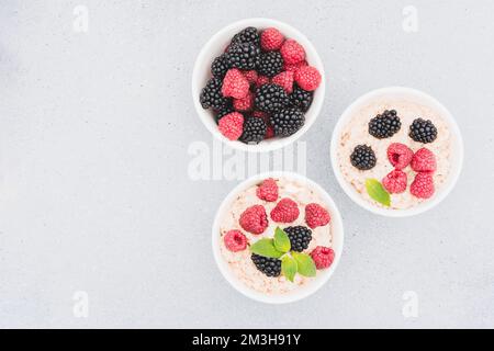 Porridge de flocons d'avoine garni de diverses baies dans des bols blancs sur fond de table en béton avec espace de copie. Recette de porridge du matin Banque D'Images