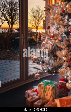 Des arbres hivernaux et une vue sur la mer pendant un coucher de soleil doré vu par une fenêtre de patio. Il y a un joli arbre de Noël dépoqué et givré avec Fair Banque D'Images