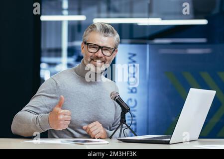 Portrait d'un beau homme senior assis dans un bureau, studio à une table avec un microphone. Enregistrez le podcast, l'entretien, l'audio. Un homme souriant regarde la caméra. Il pointe un super doigt. Banque D'Images