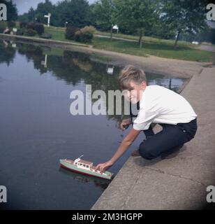 Fin '60s, historique, un garçon jouant avec son bateau modèle en bois sur l'eau, au bord d'un lac de navigation dans le parc, Angleterre, Royaume-Uni. Construire et jouer avec des bateaux modèles a été une activité populaire dans les 1950s et 1960s et le bateau vu ici pourrait être un modèle fait par Maycraft. Banque D'Images