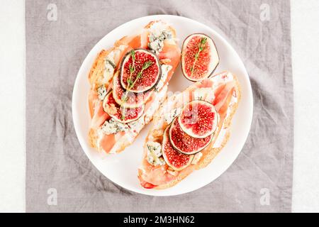 Pain grillé à la bruschetta croissant avec figue mûre, fromage bleu, prosciutto, noix et herbes sur une assiette blanche. Mise au point sélective Banque D'Images