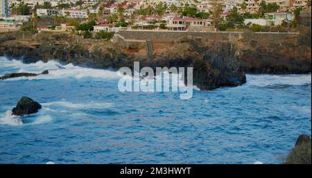 Océan de tempête et vagues éclaboussant contre les roches volcaniques. Ténérife, Îles Canaries, Espagne. Dans la rive arrière-plan de la ville de Puerto de Banque D'Images