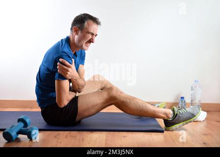 Homme faisant du sport à l'intérieur se plaignant de douleurs aux épaules assis sur un tapis. Vue latérale Banque D'Images