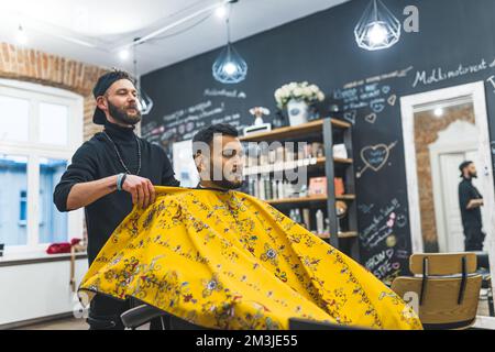 barbier professionnel mettant sur un cape à motif jaune sur son client élégant. Prise de vue moyenne en intérieur. Homme indien moyen adulte au salon de coiffure. Photo de haute qualité Banque D'Images