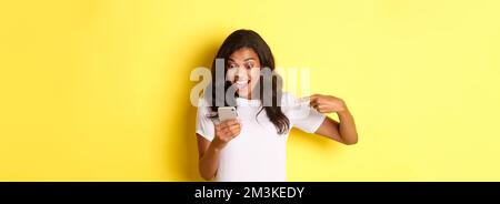 Image d'une jeune fille afro-américaine excitée, regardant et pointant du doigt sur un téléphone mobile, voyant quelque chose d'incroyable en ligne, debout sur fond jaune Banque D'Images