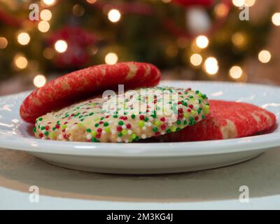 Gros plan de trois biscuits au sucre de Noël décorés de sucre rouge et de saupoudrés ronds verts, rouges et blancs sur une assiette blanche. Photographié avec le doit Banque D'Images