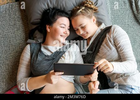 Les filles caucasiennes gaies s'étendent sur le sol et regardent des vidéos amusantes sur une tablette numérique. Bonne relation, à la maison en appréciant la journée de repos Banque D'Images