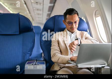 Un homme d'affaires ou un PDG asiatique prospère est assis à la fenêtre en classe affaires, en vérifiant son montre pour l'heure d'arrivée, sur le vol pour son ove Banque D'Images