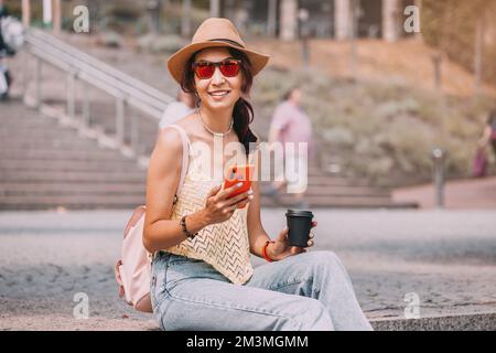 Une fille est assise sur un escalier dans le parc avec une tasse de café et lit un fil d'actualité sur un réseau social via son smartphone. Connexion Internet Wi-Fi Banque D'Images