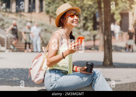 Une fille est assise sur un escalier dans le parc avec une tasse de café et lit un fil d'actualité sur un réseau social via son smartphone. Connexion Internet Wi-Fi Banque D'Images