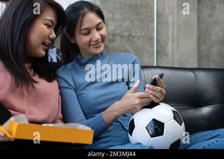 Vos amis regardent des événements sportifs sur leur téléphone mobile, applaudissent et célèbrent. Les fans de supporters asiatiques sont heureux et variés. Ils s'assoient sur le canapé avec du pop-corn et des boissons Banque D'Images