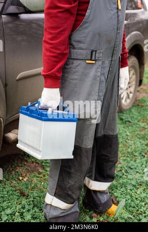 Un mécanicien de voitures en combinaison porte une batterie de voiture pour le remplacement. Charge et réparation des accumulateurs. Banque D'Images