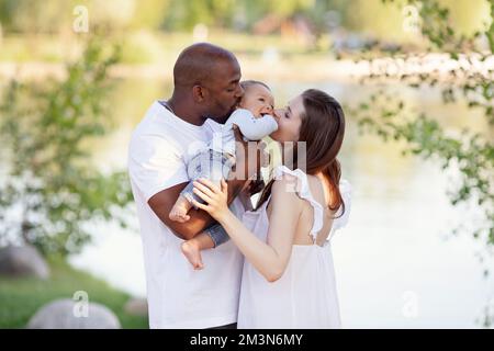 Bonne famille multiethnique. La mère caucasienne et le père afro-américain embrassent un petit enfant. Parents, Portrait de maman, papa et bébé sur les mains de la nature b Banque D'Images