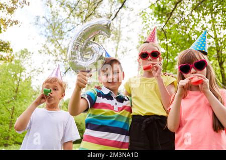 Joyeux garçons et filles avec des chapeaux de fête soufflantes soufflantes à l'anniversaire dans le jardin Banque D'Images