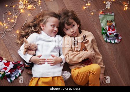 Portrait d'enfants adorables, allongé sur le sol à la maison, à l'intérieur avec décoration de Noël, guirlande d'ampoules et de chaussettes. Beau garçon et fille portent s Banque D'Images