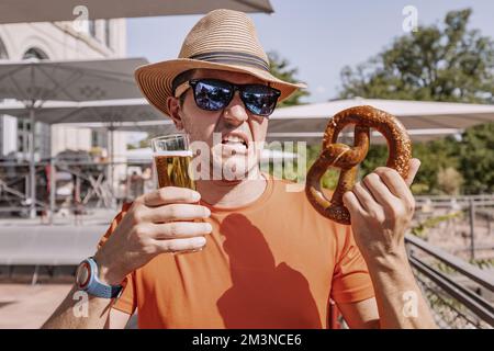 Un homme drôles boit de la bière légère fraîchement brassée à partir d'un verre et mange du bretzel dans un bar, un pub ou un biergarten allemand Banque D'Images