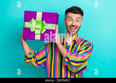 Photo de l'homme excitée par l'électricité statique impressionné brunet cheveux vêtus chemise rayée montrant boîte-cadeau avec regard isolé sur fond turquoise couleur Banque D'Images