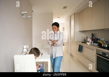Belle femme croisant les mains tout en contrôlant son fils dans la cuisine Banque D'Images