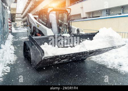 Mini-tracteur chargeuse à direction à ski nettoyer la neige sale de la voie de conduite. Nettoyage de la rue de la ville, élimination de la neige glacée à de fortes chutes de neige et de blizzard. Chasse-neige Banque D'Images