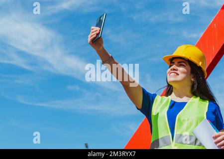 jeune femme latine caucasienne, étudiante en génie prenant un selfie avec son téléphone mobile, pendant un stage dans un chantier de construction. Banque D'Images