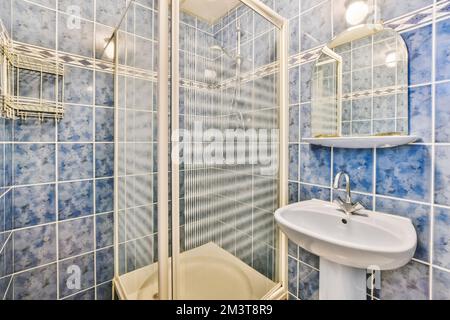 une salle de bains avec des carreaux bleus sur les murs et des accessoires blancs dans la cabine de douche est à côté d'un petit lavabo Banque D'Images