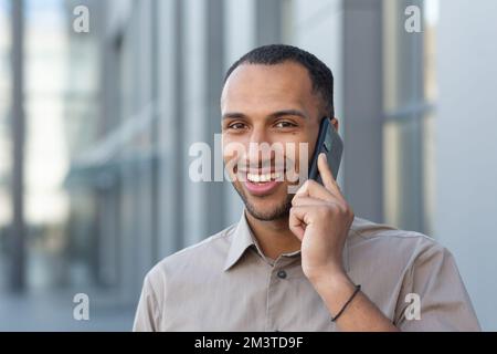 Photo en gros plan d'un homme souriant à l'extérieur d'un immeuble de bureau, qui parle au téléphone avec joie, un homme d'affaires dans une chemise décontractée hispanique regardant le portrait de l'appareil photo. Banque D'Images