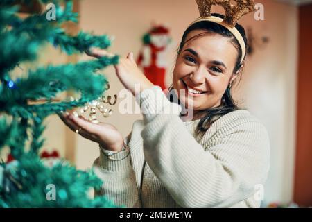 Monter l'arbre me met dans l'ambiance festive. Portrait d'une jeune femme heureuse décorant son arbre de Noël à la maison. Banque D'Images