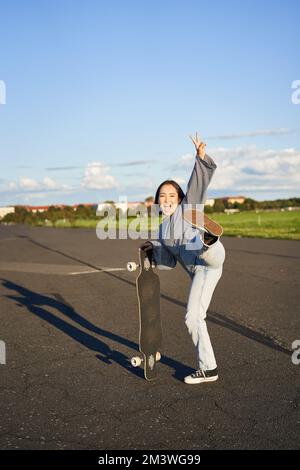 Portrait vertical d'une femme asiatique souriante debout sur la route avec la longboard, le skateboard sur une longue croiseur, posant sur une route vide par beau temps Banque D'Images