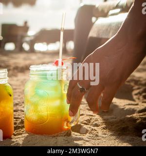 Une main féminine tenant un cocktail coloré Michelada sur une plage de sable Banque D'Images