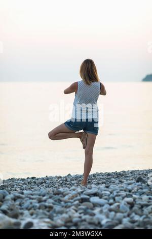 Une femme menant un mode de vie sain et pratiquant le yoga, exécute vrikshasana exercice sur la mer, pose d'arbre, se tient au coucher du soleil contre le dos Banque D'Images