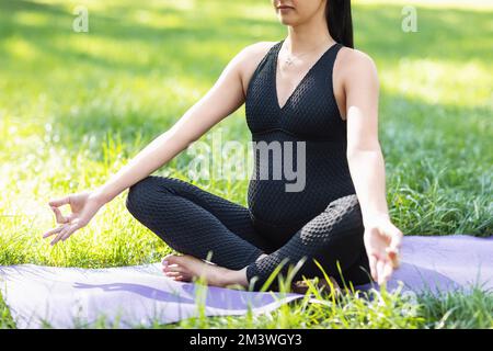 Une jeune femme enceinte médite assise dans une position de lotus avec un symbole de mudra jnana, s'entraîne dans une combinaison de sport noire dans un parc sur l'herbe, prac Banque D'Images