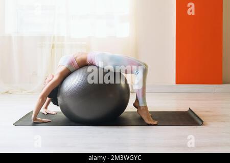 Une femme attirante menant un mode de vie sain et pratiquant le yoga, exécute une variante de l'exercice Urdhva Dhanurasan avec une balle de fitness, un bri Banque D'Images
