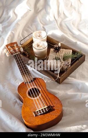 Noël matin confortable à la maison. Boîte cadeau et aubergine sur plateau en bois près de la guitare ukulele sur le lit. Banque D'Images