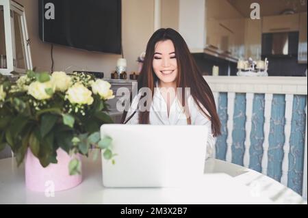 Jeune femme d'affaires asiatique belle travaillant en ligne avec ordinateur portable placé à la table au bureau à domicile. Femme souriante freelance ont le travail à distance assis dans la salle de séjour avec des fleurs roses blanches Banque D'Images