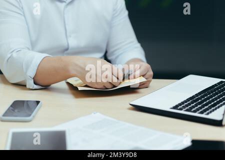 Gros plan. Les mains d'un homme à une table avec un ordinateur portable et les documents prennent de l'argent d'une enveloppe, compte l'argent. Banque D'Images