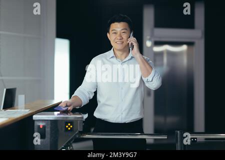 Un jeune homme asiatique, le directeur, homme d'affaires, entre dans le bureau par un tourniquet électronique, un système de sécurité automatique. Contient sa carte d'accès électronique. Parler au téléphone, sourire. Banque D'Images