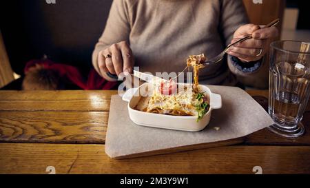 Une femme mange de la lasagne avec du fromage et une sauce Behamel à partir d'une plaque en céramique blanche située à côté d'un verre d'eau sur la table Banque D'Images