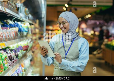 Femme vendeur dans le super marché dans hijab avec des produits de vérification de tablette en utilisant un ordinateur de poche, femme musulmane près des étagères avec des produits et des marchandises. Banque D'Images