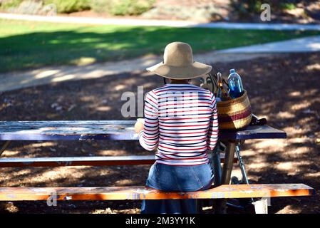 une femme assise sur un banc de parc portant un chapeau de disquettes en fibre naturelle a un déjeuner paisible et calme dans un cadre semblable à celui d'un parc Banque D'Images