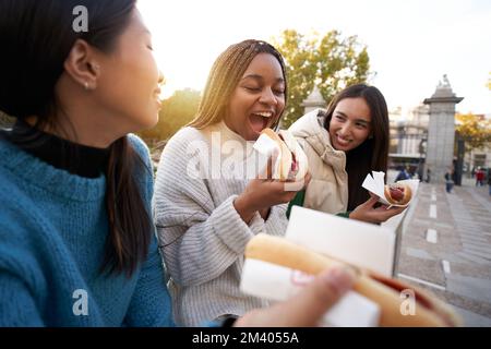 Gaiful Girls Eating Take Away Street Food. Trois amies heureuses qui prennent des hot dogs dans le parc Banque D'Images