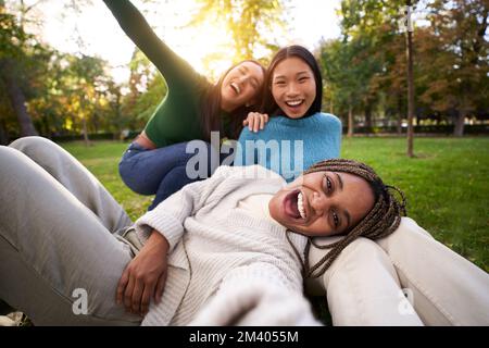 Trois filles multiraciales qui se trouvent sur l'herbe prennent le selfie en regardant l'appareil photo. Groupe d'amis féminins Banque D'Images