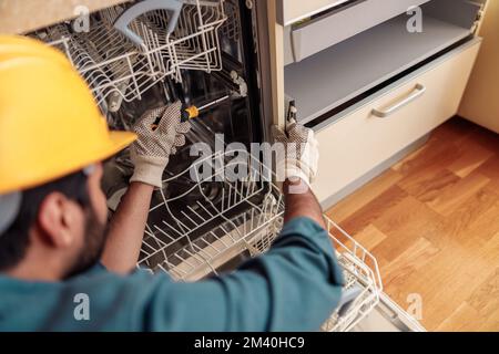 Gros plan de handyman dans des vêtements spéciaux de réparation lave-vaisselle dans la cuisine moderne Banque D'Images