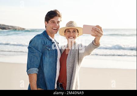 Certains des meilleurs souvenirs sont faits à la plage. un couple d'âge moyen prenant un selfie sur la plage. Banque D'Images