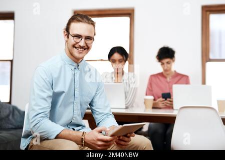 Cette équipe fait les choses. Portrait court d'un beau jeune homme d'affaires assis et utilisant une tablette pendant que ses collègues travaillent derrière lui. Banque D'Images