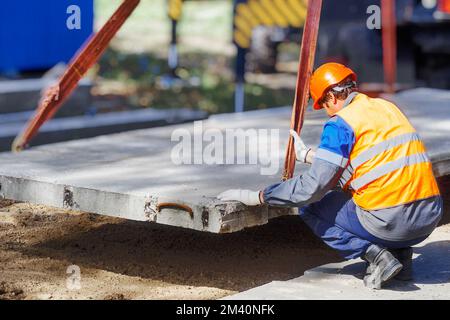 Le Slinger pose une dalle de béton sur le chantier de construction le jour de l'été. Le travailleur dans la veste de protection et le casque de construction supervise la pose de la base sur le chantier. Banque D'Images