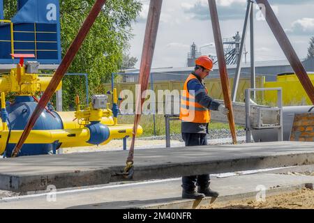 Le Slinger pose une dalle de béton sur le chantier de construction le jour de l'été. Le travailleur dans la veste de protection et le casque de construction supervise la pose de la base sur le chantier. Banque D'Images