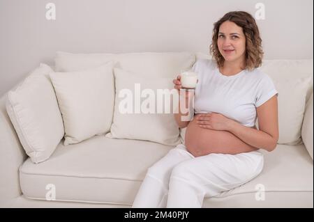 Une femme enceinte s'assoit sur un canapé blanc et boit du lait. Banque D'Images