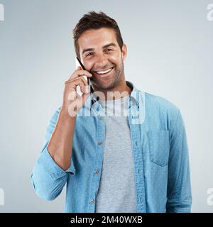 C'est vraiment génial d'entendre parler de vous. Portrait d'un jeune homme joyeux parlant sur son téléphone portable tout en se tenant sur un fond gris. Banque D'Images