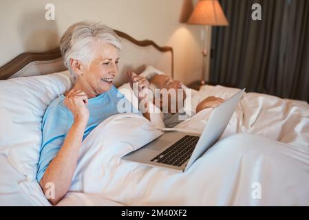 Oh, j'ai hâte de lui montrer cela. une femme âgée assise au lit et utilisant un ordinateur portable pendant que son mari dort à côté d'elle. Banque D'Images